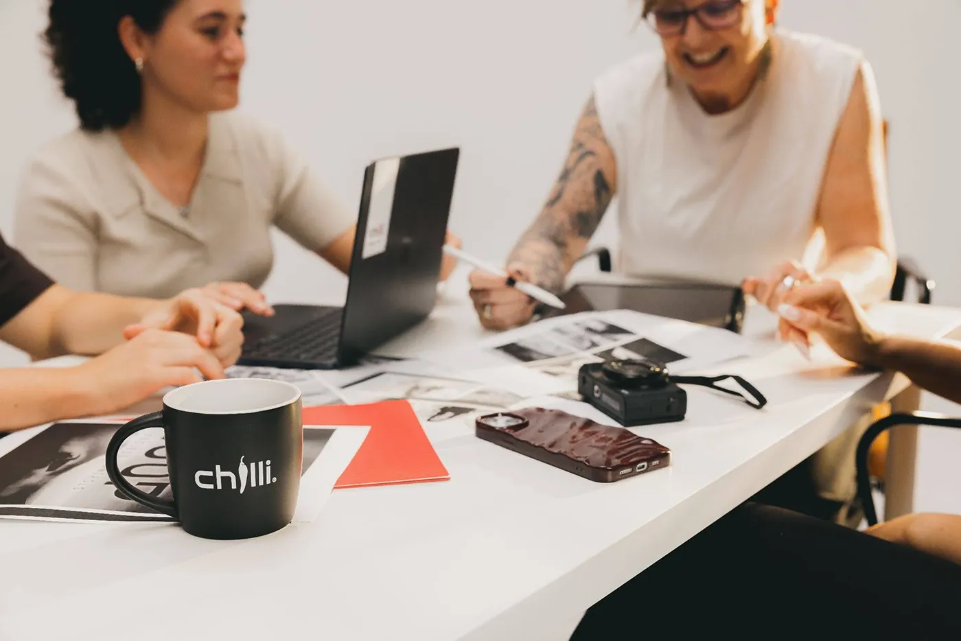 Hospitality & Food - Chilli Black coffee mug with chilli digital marketing agency logo on the mug on a table with people sitting around looking at pieces of paper, laptop, camera, and phone.
